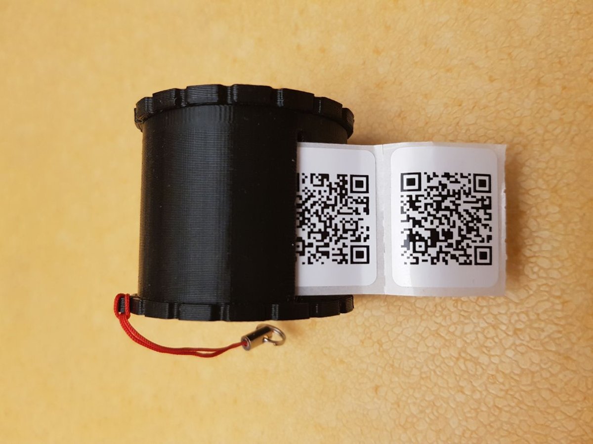 Fekete, hengeres QR-kód tartó fekvő helyzetben, két QR-kódos címke lóg ki belőle, piros zsinóros akasztóval