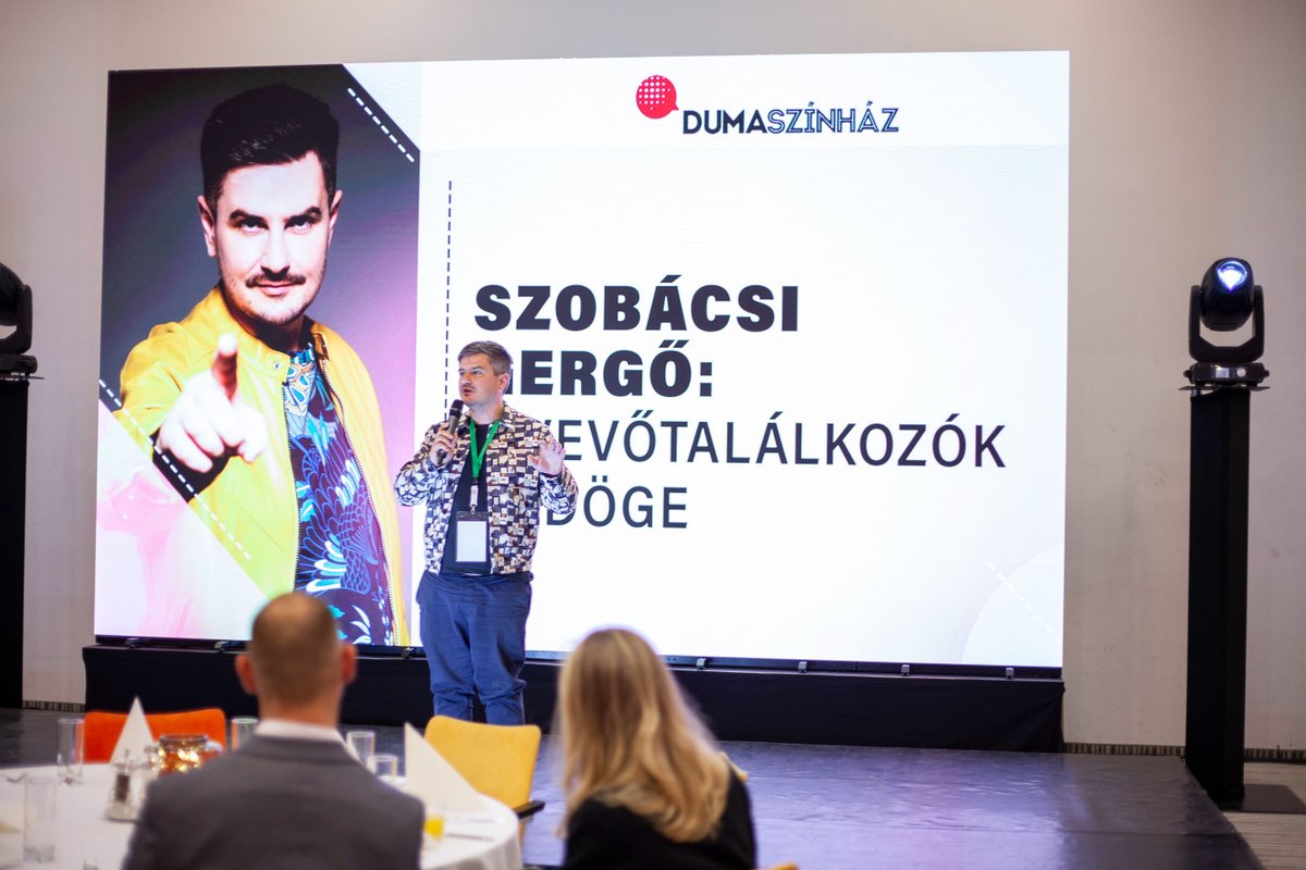 Előadó mikrofonnal a színpadon, mögötte a Dumaszínház kivetítője Szobácsi Gergő nevével, előtérben a közönség