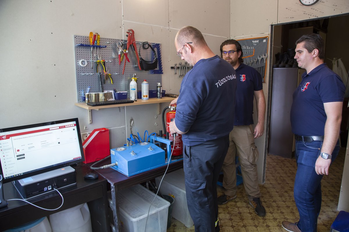 Tűzoltóság feliratú pulóveres szerelő tűzoltó készülék nyomáspróbáját végzi a műhelyben, a monitoron a fiREG felülete