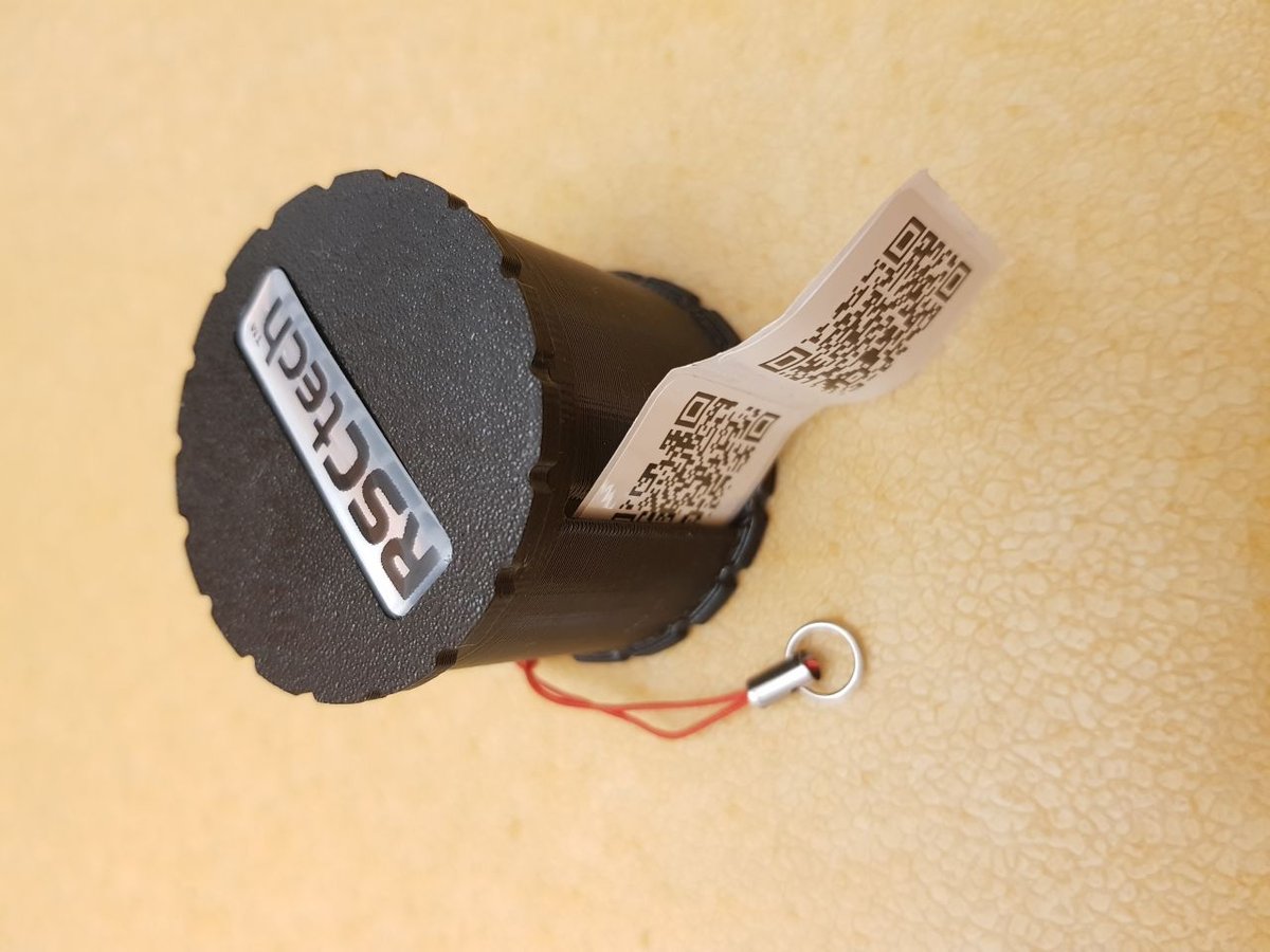 Fekete, hengeres RSCtech QR-kód tartó, amelyből QR-kódos címkeszalag lóg ki, piros zsinórral